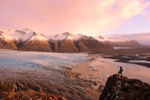 Hiker is standing at the edge of a cliff overlooking the Skaftafell Glacier in Skaftafell National Park in Iceland. The traveller is distant and looks very small in this majestic landscape on top of a mountain with the beautiful view over one of the many tongues of Vatnajokull glacier and the adjacent lagoon.